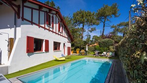 Une piscine avec une terrasse en bois, un bâtiment blanc agrémenté de bordures rouges et une pelouse bien entretenue.