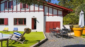 Une maison à deux étages avec une porte et des fenêtres rouges, une terrasse avec une table et des chaises, et un petit espace piscine.