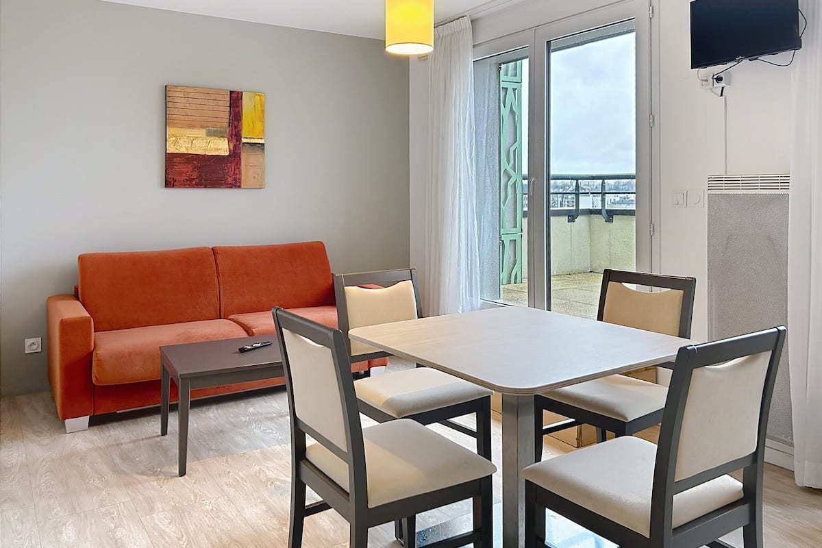 A modern living room with a sofa, a dining table with chairs, a television mounted on the wall, and a large window with curtains.