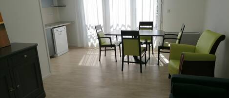 A modern living room with a dining area, a kitchen, and a view through sheer curtains.