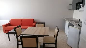 A compact living space with a dining table, four chairs, a red sofa, a kitchenette with a microwave, and a small refrigerator.