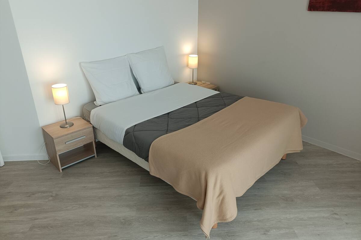 A neatly made bed with a grey quilt and a beige throw blanket, flanked by two bedside tables with lamps.