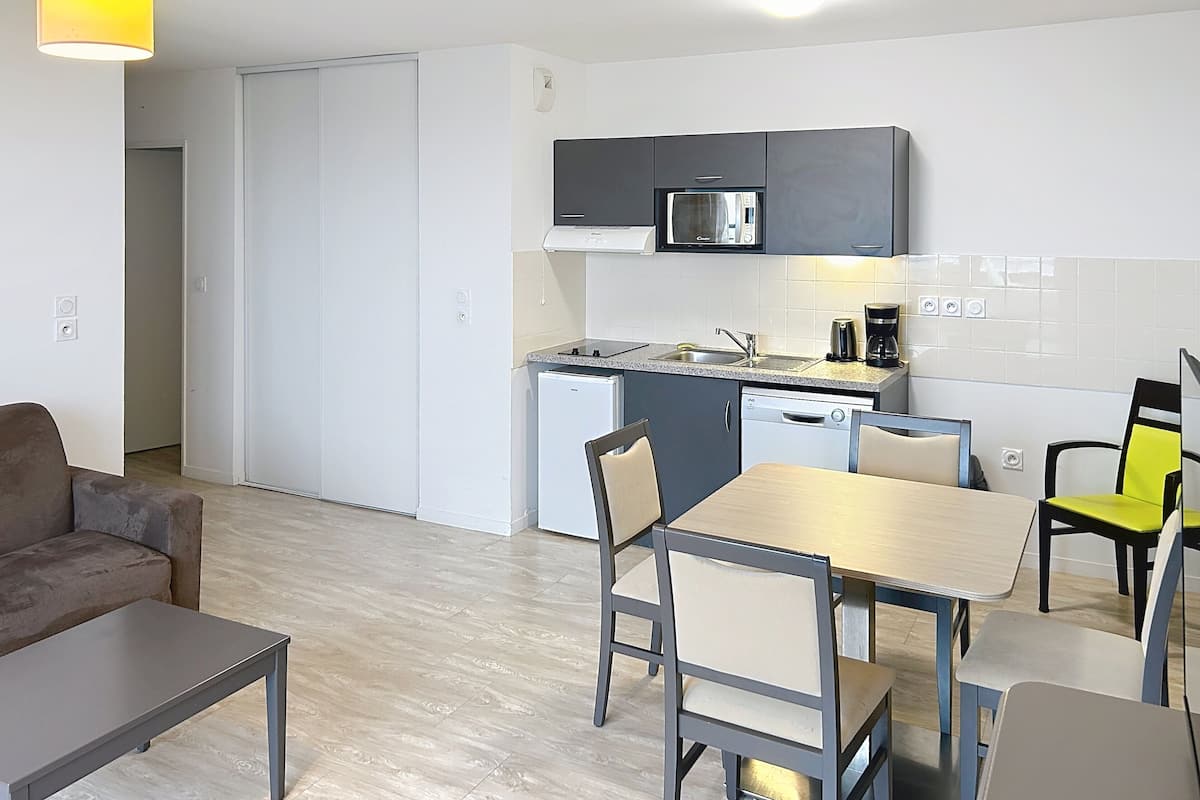 A compact kitchen with a microwave, coffee maker, and dishwasher. There is a dining table with chairs and a sofa in the living area.