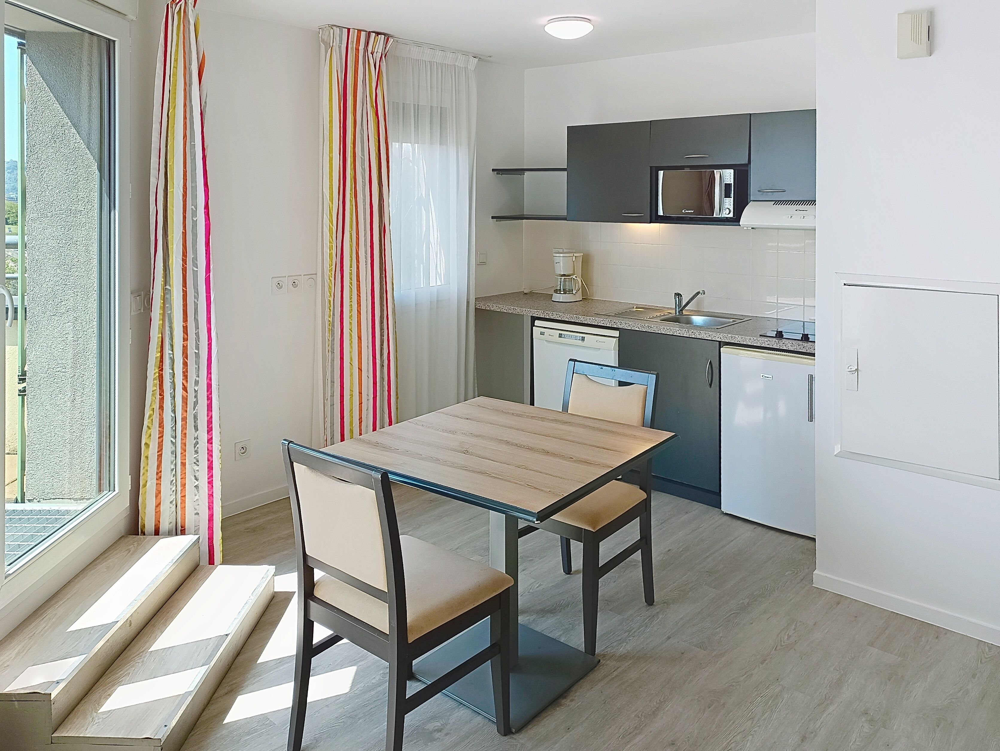 A compact kitchen with a microwave, coffee maker, and dishwasher. There is a dining table with two chairs and a window with curtains.