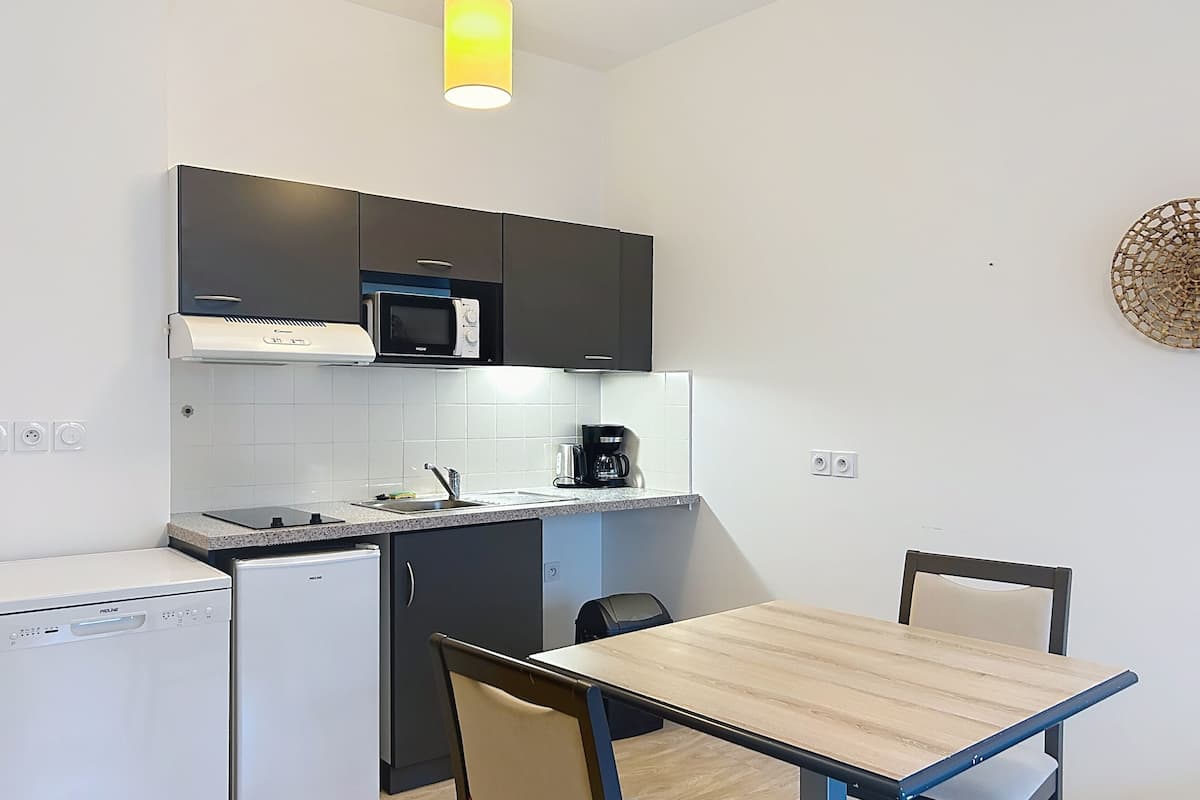 A compact kitchen with a microwave, coffee maker, and a small refrigerator. There is a dining table with chairs and a hanging light fixture above.