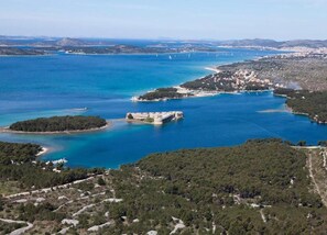 Vista aerea di una zona costiera con una struttura fortificata circondata da acqua, vegetazione fitta e un paese in lontananza.
