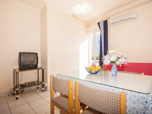 Een eetgedeelte met een glazen tafel, stoelen, een televisie op een standaard, een fruitschaal en een vaas met bloemen.