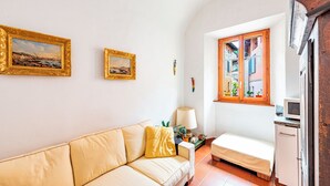 Un espace de vie compact comprenant un canapé, une petite table avec une lampe, un lit et une fenêtre donnant sur des immeubles.