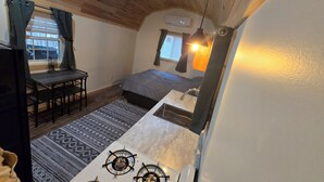 Une petite pièce comprenant un lit, une petite table, une chaise, un réchaud et une fenêtre avec des rideaux.