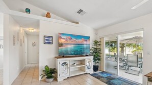 Una sala de estar moderna con un televisor grande, una puerta de vidrio que da a un área de estar exterior y una planta en maceta.