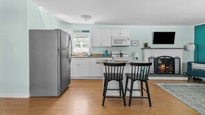 Une cuisine moderne avec des armoires blanches, un réfrigérateur en acier inoxydable, un four à micro-ondes et une cuisinière à gaz. On y trouve un coin repas avec deux chaises noires et une petite table. Un téléviseur à écran plat est fixé au-dessus de la cheminée.