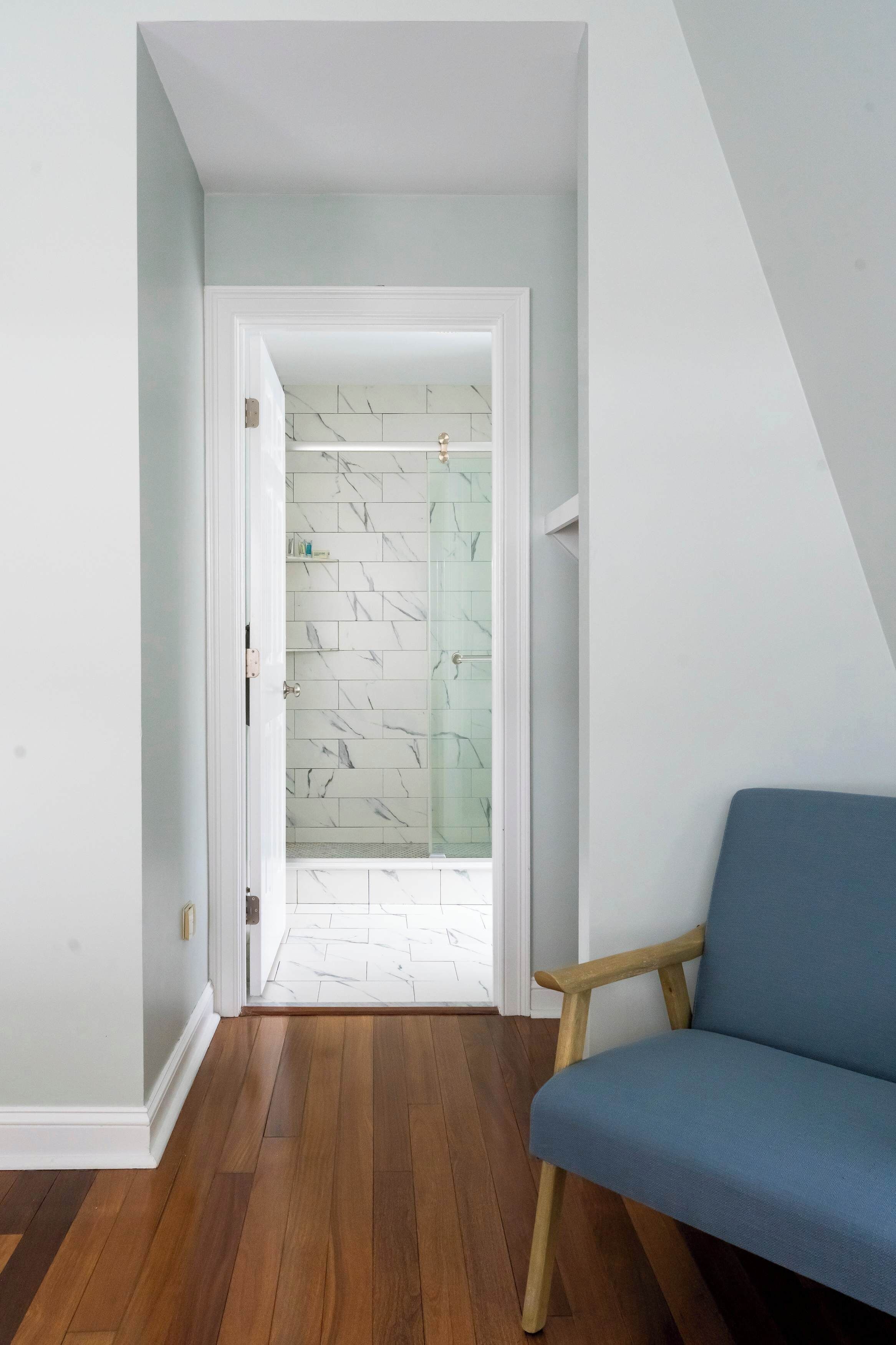 A room with a wooden floor, a blue chair, and a glass shower door with white tiles.