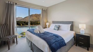 A hotel room with a large bed, two bedside tables, a chair, and a view of a lake and mountains.