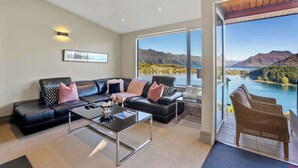 A modern living room with a black leather sofa, a glass coffee table, and a view of a lake and mountains through large windows.