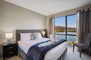 Une chambre d’hôtel avec un grand lit, une table de chevet avec une lampe, une chaise et une vue sur un lac par une grande fenêtre.
