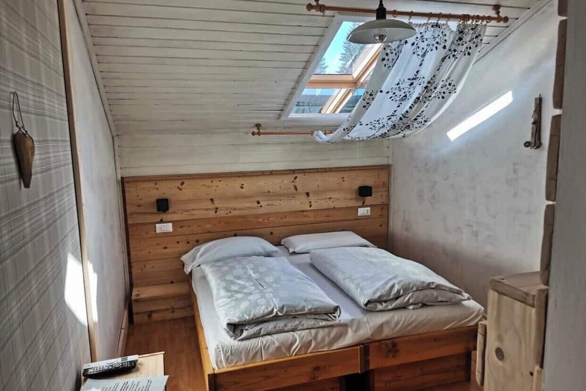 A small, cozy bedroom with a wooden headboard, a single bed with two pillows, a bedside table with a lamp, and a small window with a curtain.