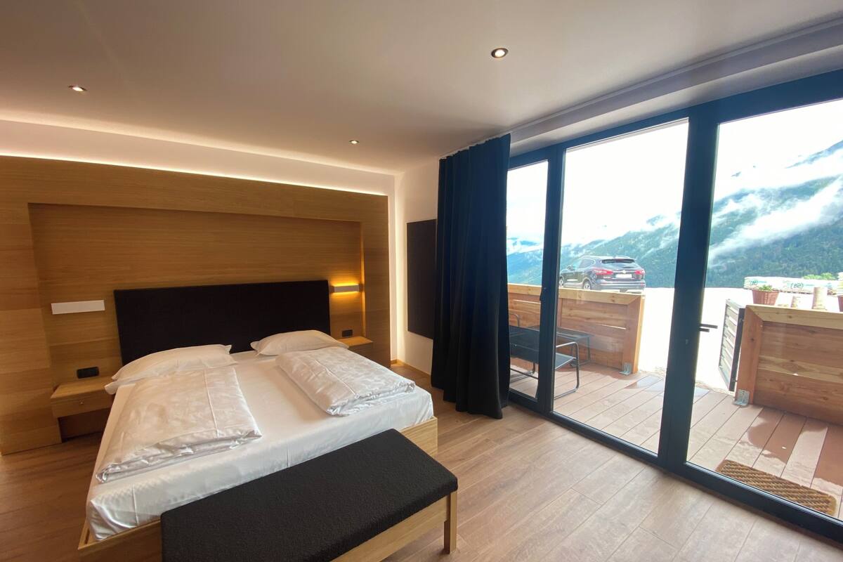 A modern hotel room with a large bed, a bench, a wooden headboard, a nightstand, and a view of a mountain landscape through a sliding glass door.