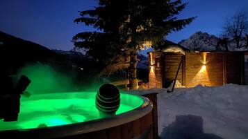 Una bañera de madera con luz verde, rodeada de nieve y una cabaña de madera con luz a la derecha.