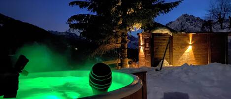 Una bañera de madera con luz verde, rodeada de nieve y una cabaña de madera con luz a la derecha.