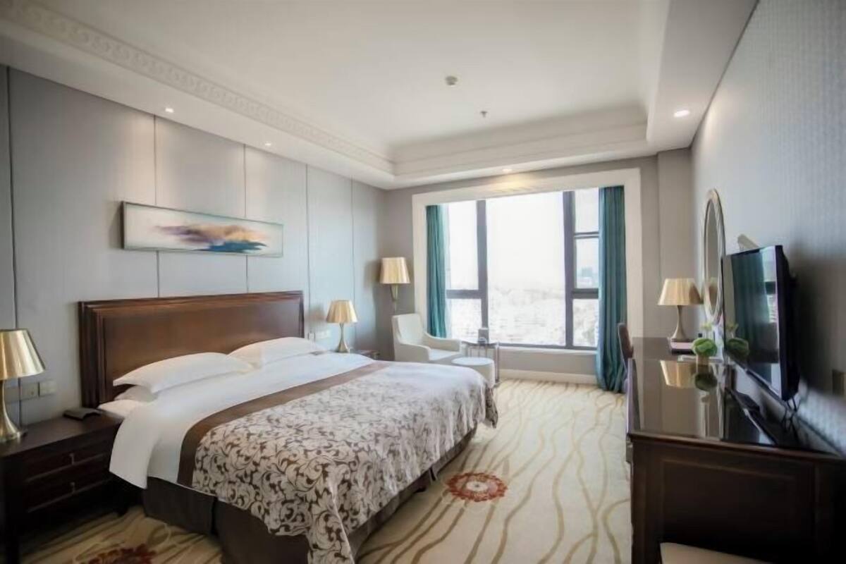 A hotel room with a large bed, two bedside tables with lamps, a TV, a chair, and a window with curtains.