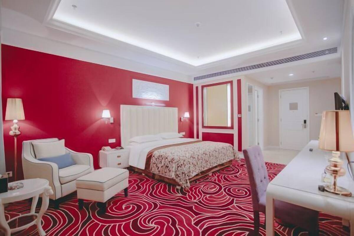 A hotel room with a red accent wall, a bed with white bedding, a white armchair, a small table, and a red patterned carpet.