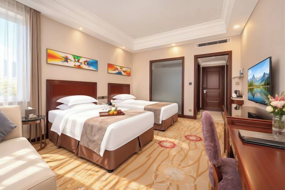 A hotel room with two beds, a desk, a TV, and artwork on the walls.