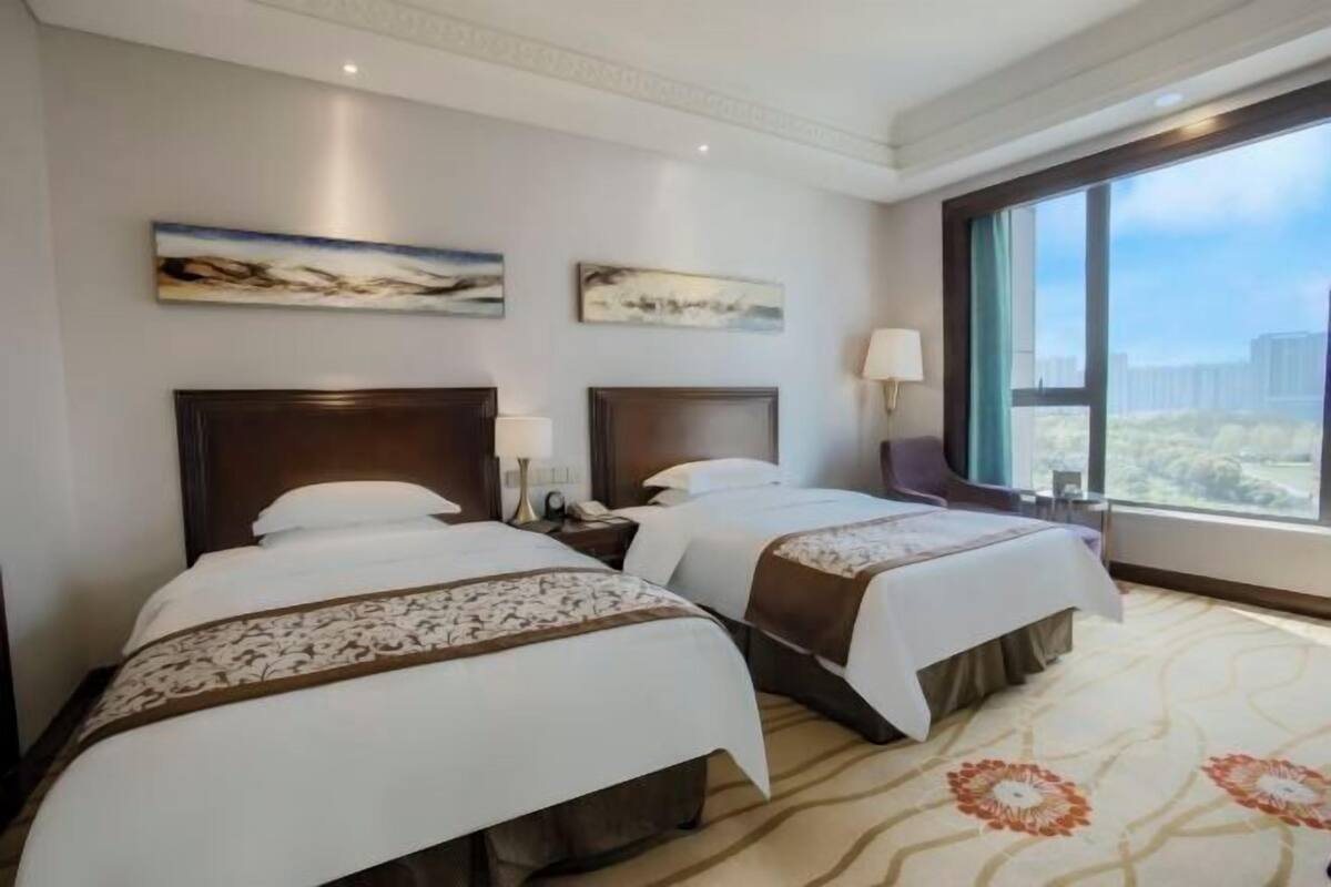 A hotel room with two beds, a large window with a city view, and artwork on the walls.