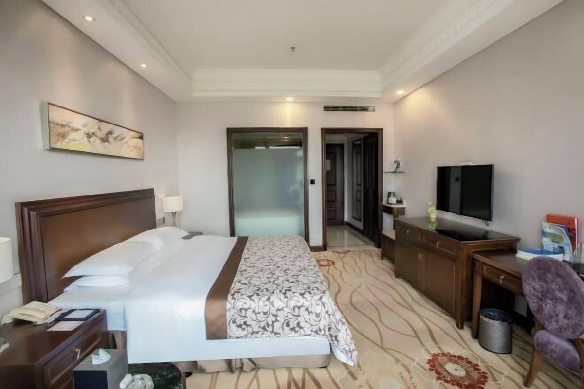 A hotel room with a bed, a desk with a television, a chair, a waste bin, and a door leading to another room.