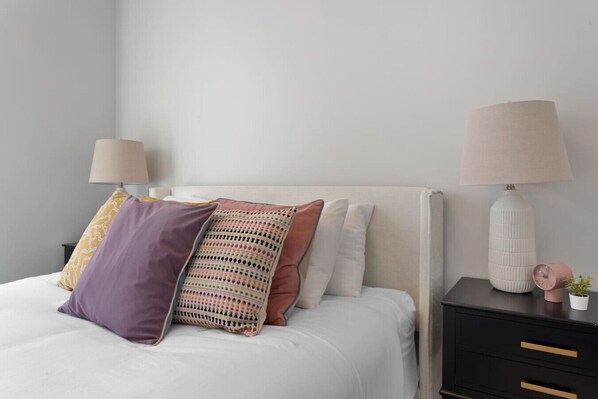 A neatly made bed with a white duvet, multiple pillows in various colors, and a bedside table with a lamp and a small potted plant.