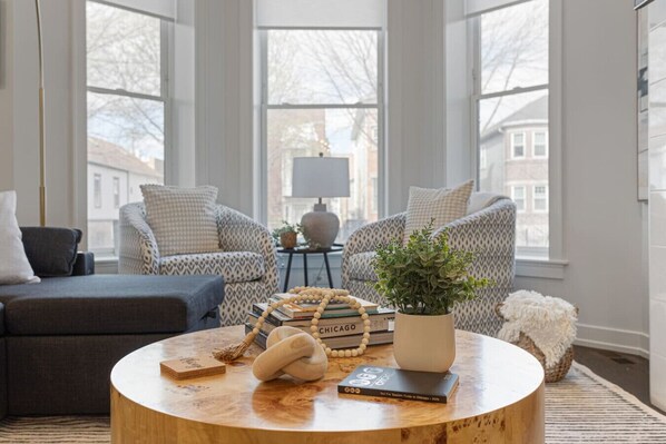 A cozy living room with a round wooden coffee table, two patterned armchairs, a dark sofa, and a potted plant.