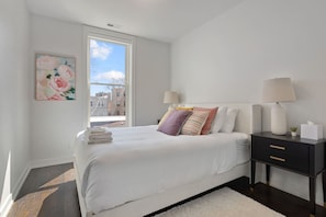 A modern bedroom with a large bed, two bedside tables with lamps, a window with a view of buildings, and a framed floral artwork on the wall. - Modern 4 Bed, 3.5 Bath Home | Near Sox & McCormick (Chicago)