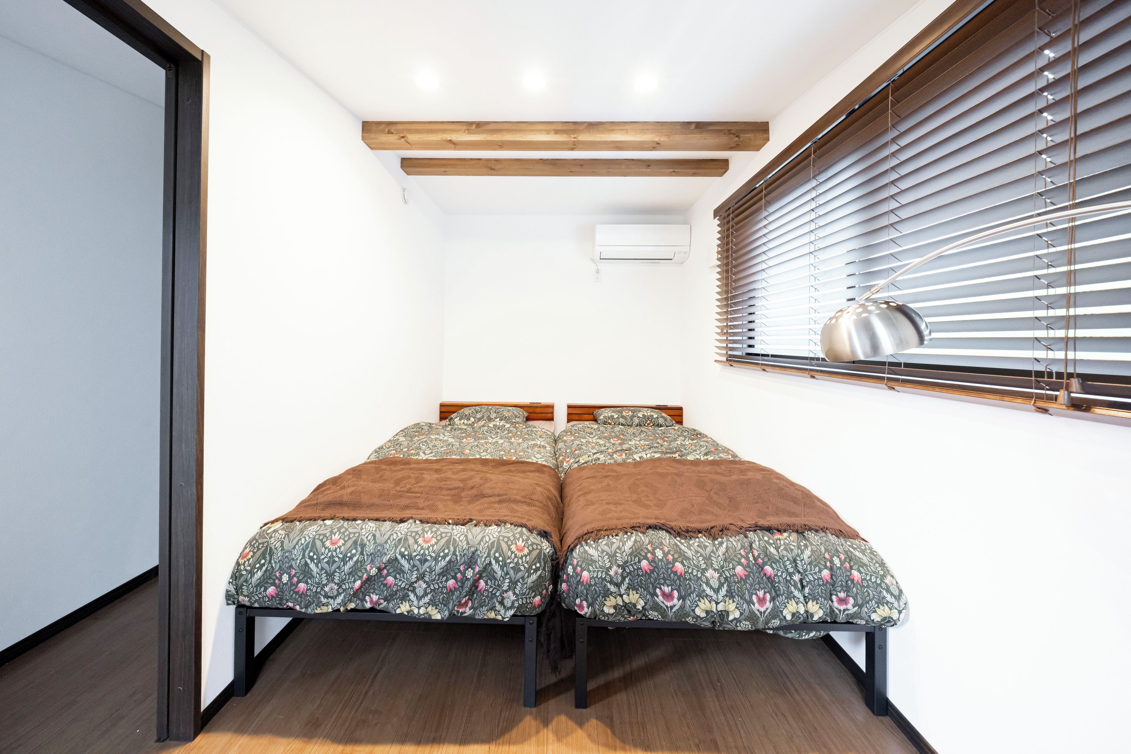 A bedroom with two beds, wooden beams on the ceiling, a window with blinds, and an air conditioning unit.