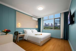 Une chambre d’hôtel équipée d’un lit, d’un bureau, d’une chaise, d’un panier de fruits, d’une fenêtre donnant sur la ville et d’un appareil de climatisation.