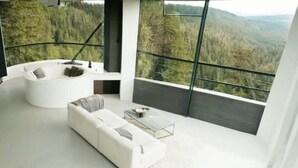 A modern living room with a white sofa, a coffee table, and a large window offering a forest view.