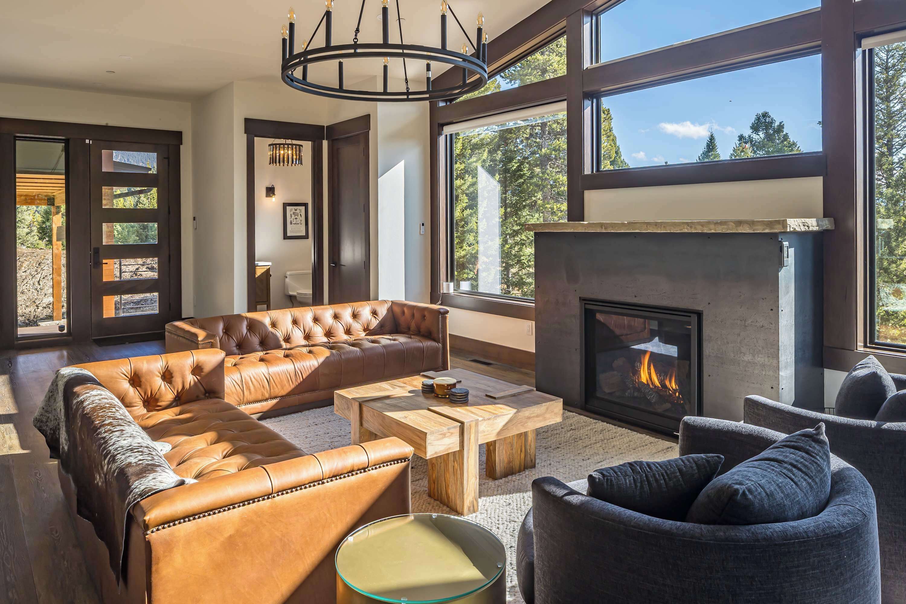 A spacious living room with a leather sectional sofa, a fireplace, and large windows offering a view of trees.