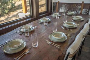 Une table à manger est dressée avec des assiettes, des verres et des couverts, et est placée près d’une fenêtre donnant sur des arbres.