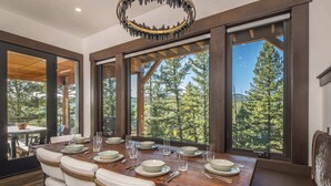 Un comedor con una gran mesa de madera dispuesta para cuatro comensales, ventanas amplias que dan a vistas de árboles y una lámpara moderna.