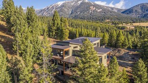 Una casa moderna de dos pisos con un amplio balcón, rodeada de un bosque denso y montañas al fondo.