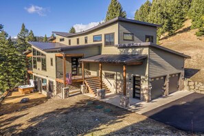 A modern two-story house with a covered porch, stone foundation, and a view of a forested hillside.