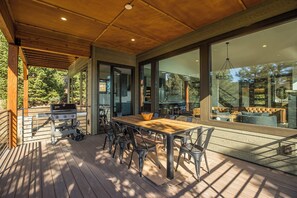 A covered outdoor dining area with a wooden deck, metal chairs, and a grill.