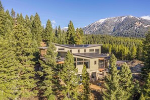 A modern two-story house surrounded by dense forest with a mountain in the background.