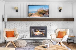 Een woonkamer met een open haard, twee fauteuils, een ronde salontafel en een schilderij met een strandthema erboven.