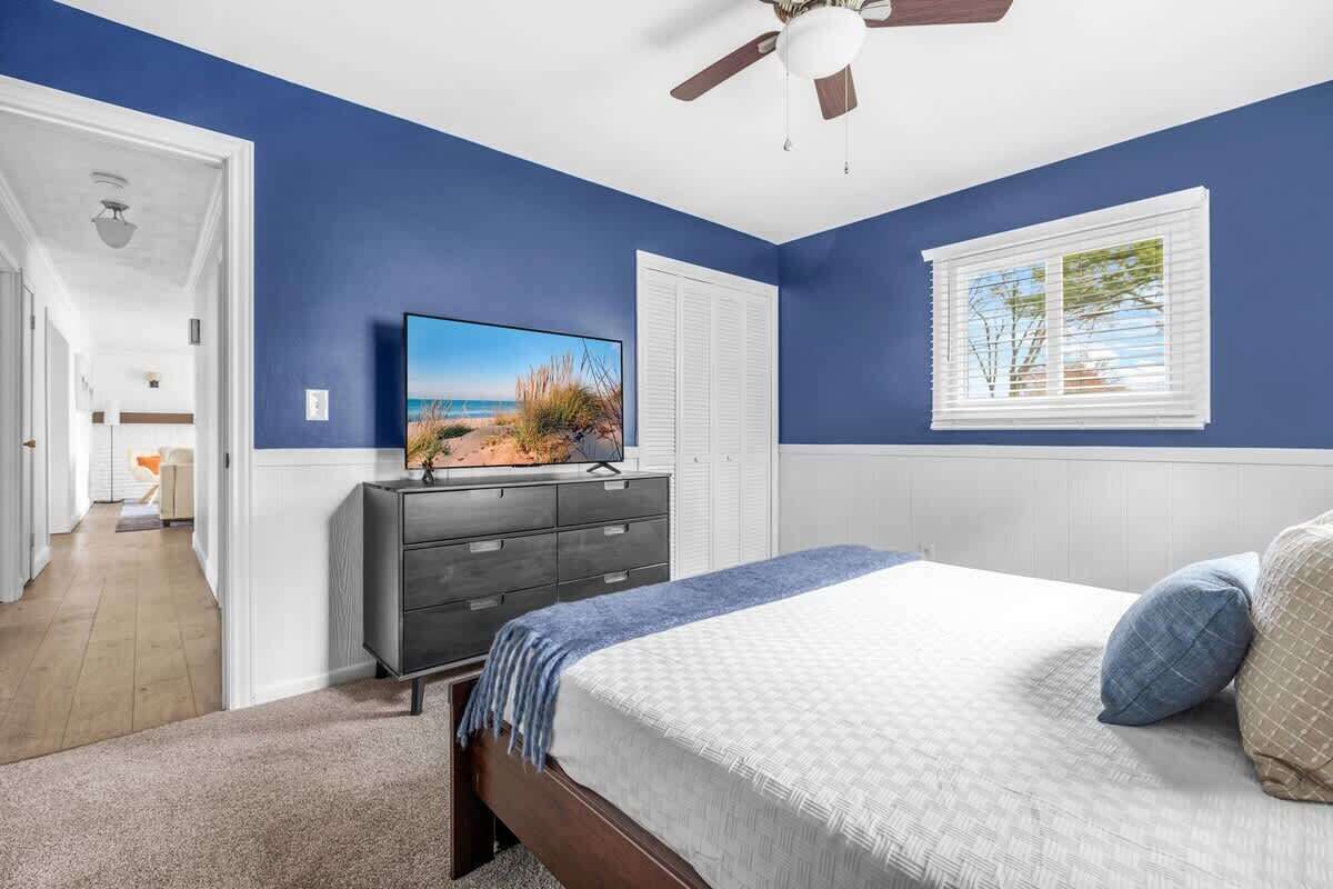 Une chambre à coucher comprenant un lit, une commode, un téléviseur et une fenêtre avec des stores.