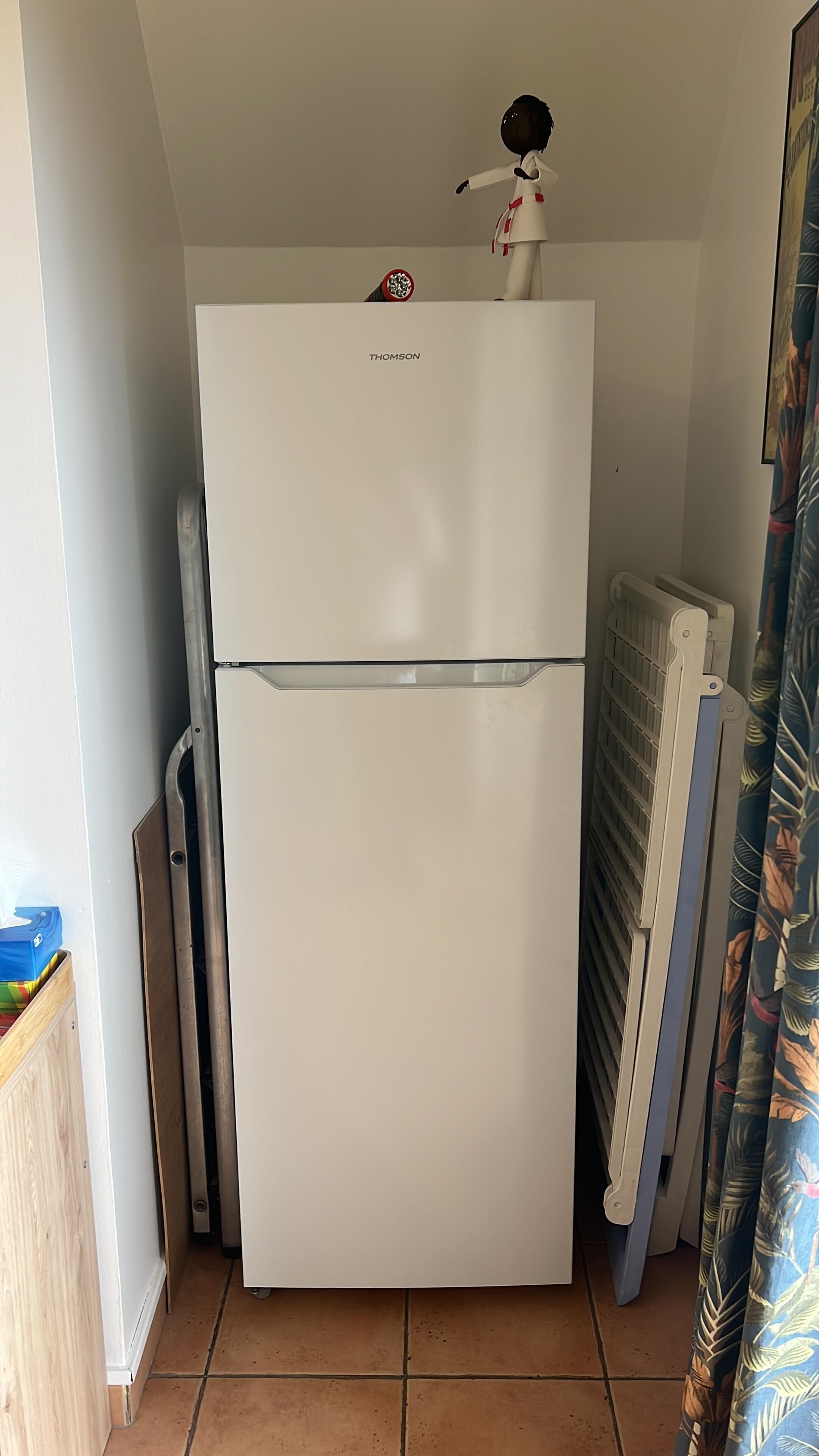 Un refrigerador Thomson blanco en una cocina con suelo de baldosas, un radiador y una cortina.