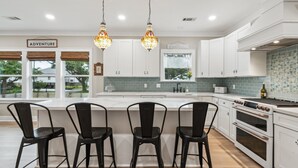 A modern kitchen with white cabinets, a large island with bar stools, and a view of the outdoors through large windows.