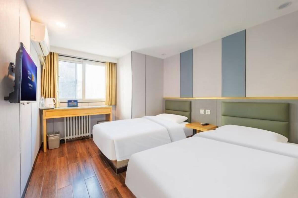 A hotel room with two single beds, a wooden floor, a window with curtains, a small table, and a TV mounted on the wall.