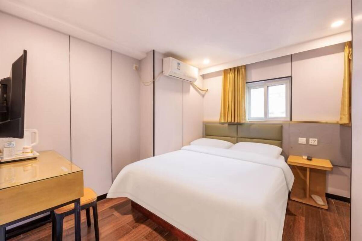 A hotel room with a bed, a desk with a chair, a television, an air conditioning unit, and a window with curtains.