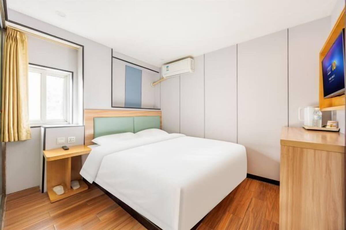 A hotel room with a bed, a desk, a chair, a TV, and an air conditioning unit.