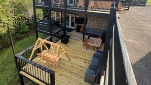 Ein zweistöckiges Gebäude mit einer Holzdachterrasse, Sitzgelegenheiten im Freien und einem großen Fenster im Obergeschoss.