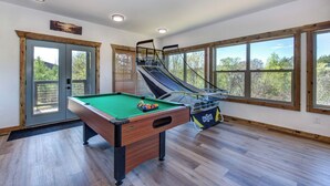 Ein Billardtisch mit grüner Spielvliesdecke, ein Billardqueue und eine Balltasche. Im Hintergrund ist eine Rutsche zu sehen, und der Raum hat große Fenster mit Blick auf Bäume.